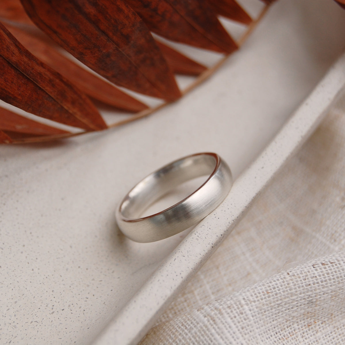 A simple matte silver band ring resting on a textured beige ceramic tray with a woven fabric and a dried brown leaf in the background.
