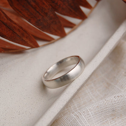 A simple matte silver band ring resting on a textured beige ceramic tray with a woven fabric and a dried brown leaf in the background.